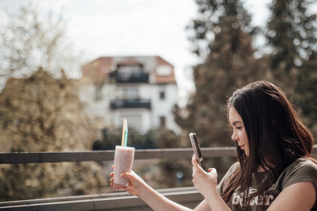 Candid authentic zoomer girl drinks freshly prepare smoothie at balcony using mobile phone outdoor